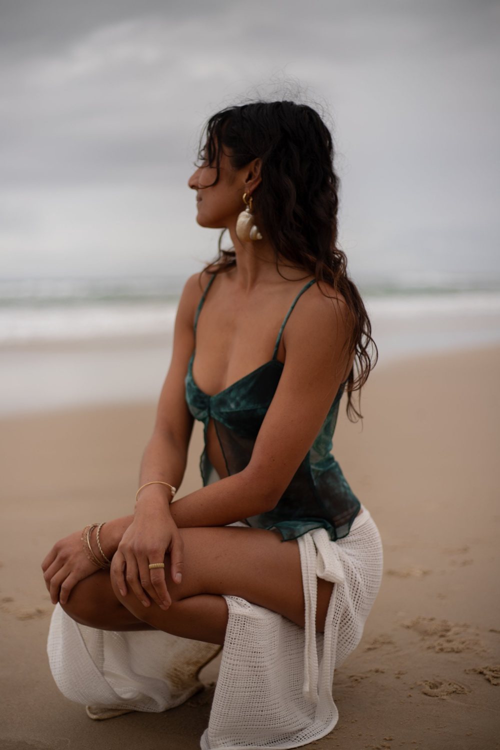 A woman crouching on the sand in a sheer green top and white crochet skirt, gazing out to sea under an overcast sky