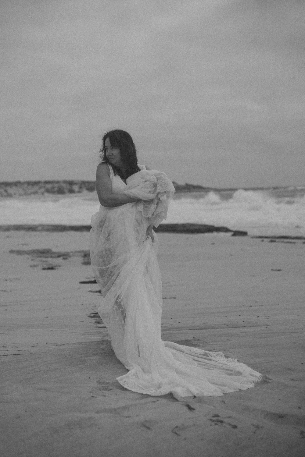 Black and white portrait of a woman on the beach wrapped in a flowing lace veil, waves breaking behind her