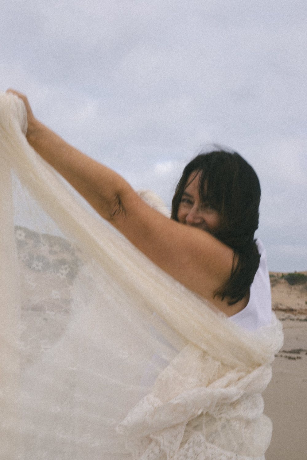 A woman laughing as she lifts a cream lace veil into the wind on the dunes
