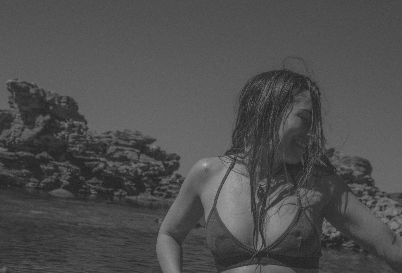 Black and white portrait of a woman laughing with wet hair across her face, rocky coastline behind her