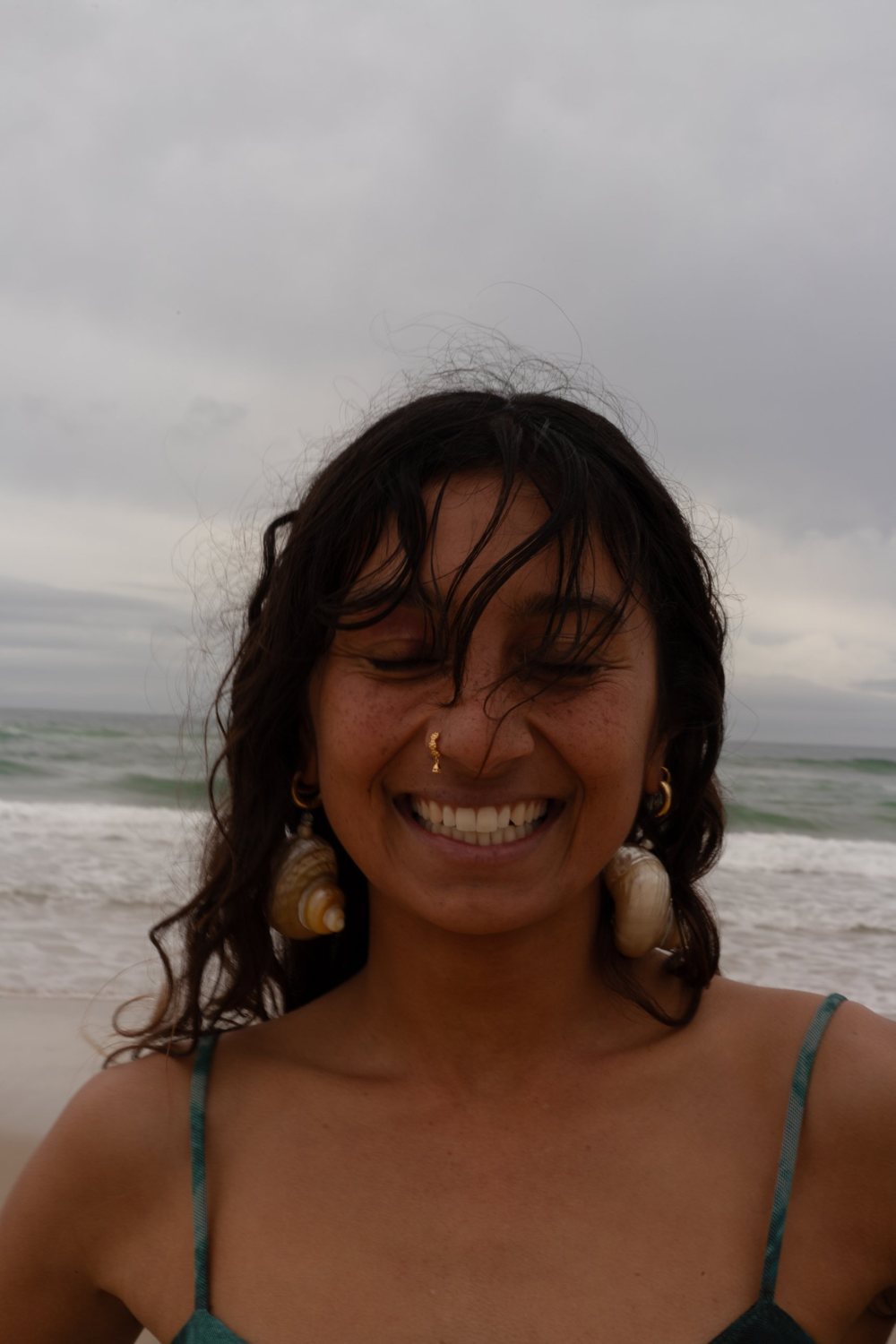 Close portrait of a woman laughing with eyes closed, wet hair and shell earrings, waves behind her