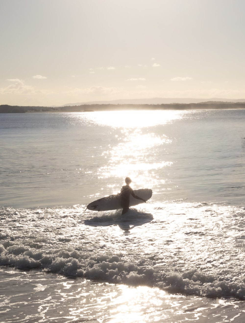 A surfer carrying a board into the shallows, backlit by sunlight sparkling across the water