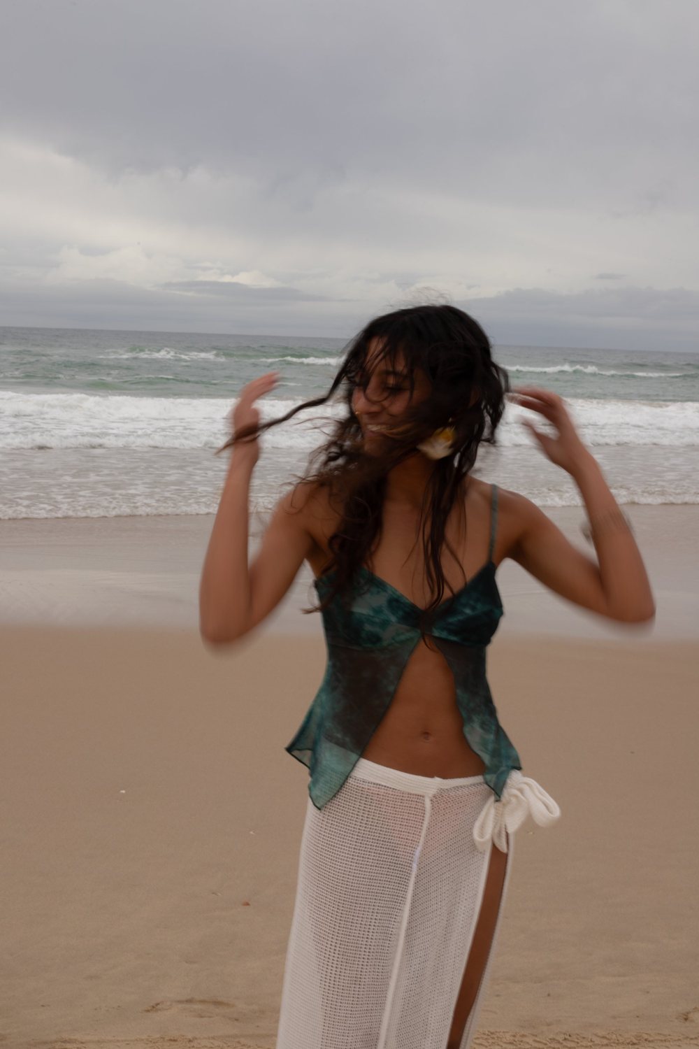 A woman spinning on the beach with her hair caught mid-movement, sheer green top and white crochet skirt