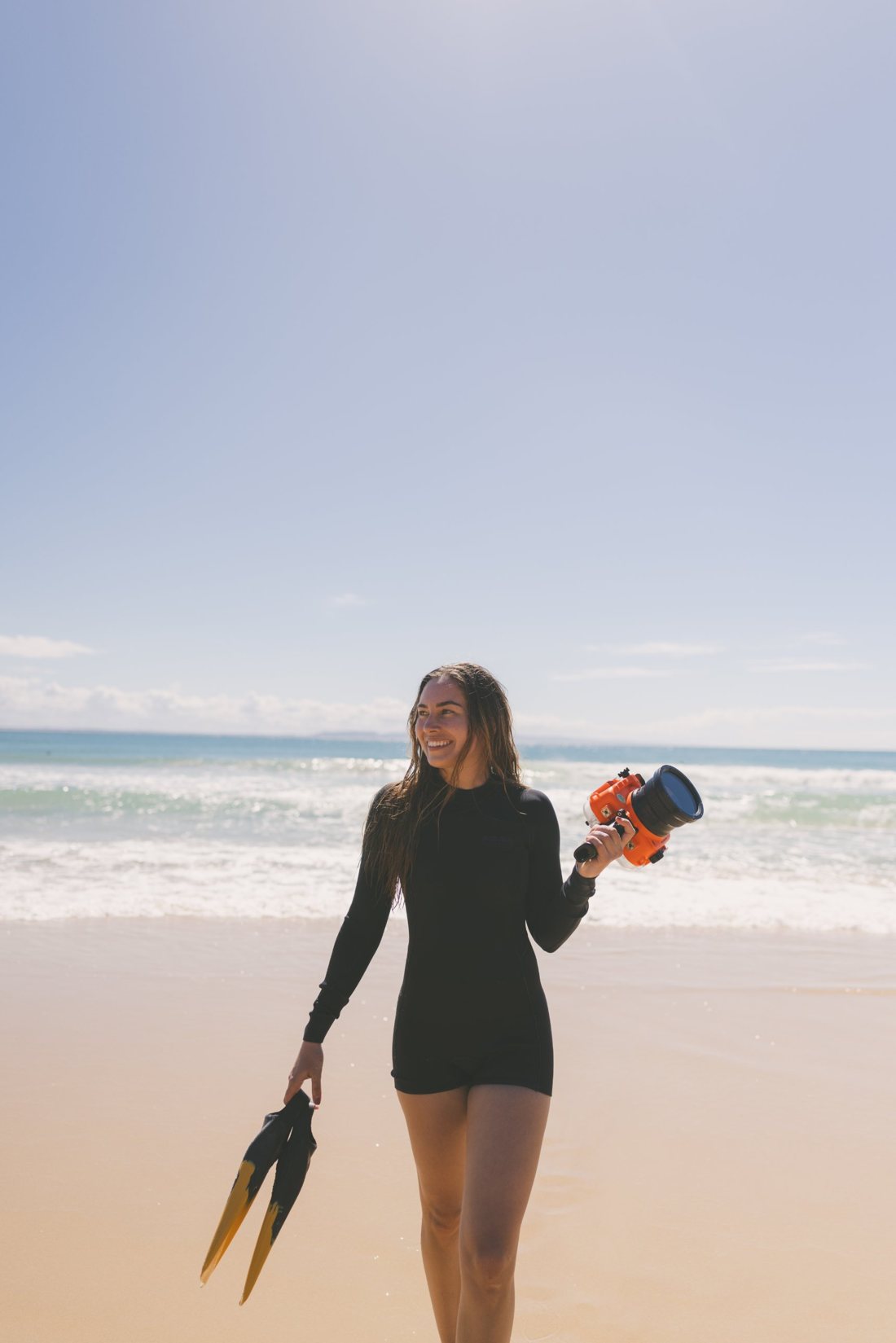 Stacey on the beach in a wetsuit, holding fins and a camera in an orange underwater housing, smiling with the surf behind her